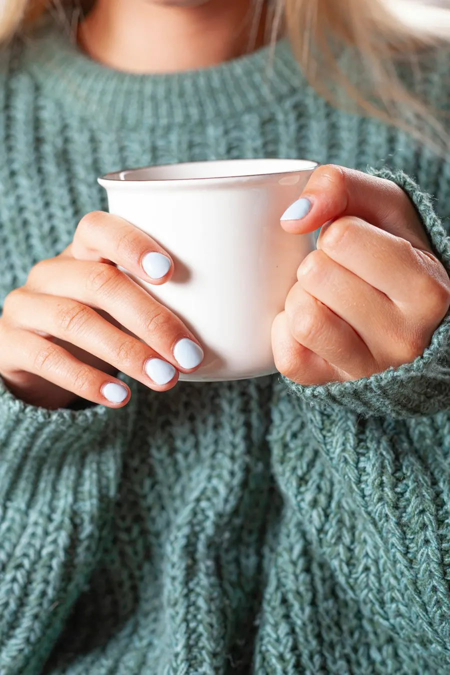 Deux mains autour d'une tasse blanche, dans un pull en maille bleu-vert